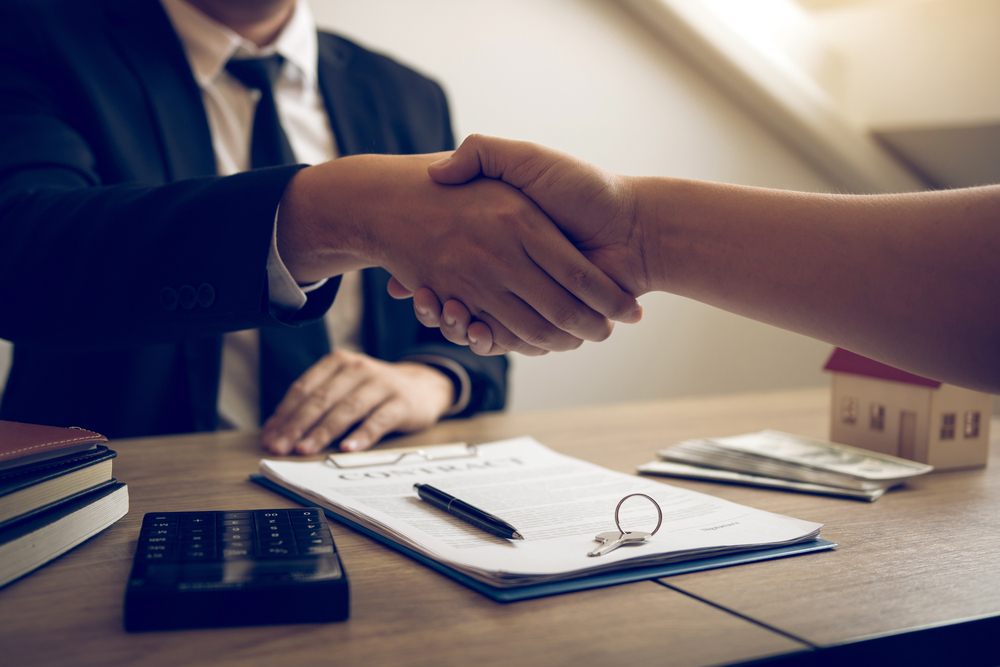 Business professionals shaking hands over a contract on a desk, with a calculator, house model, and cash visible, symbolizing agreement and data processing in market research.
