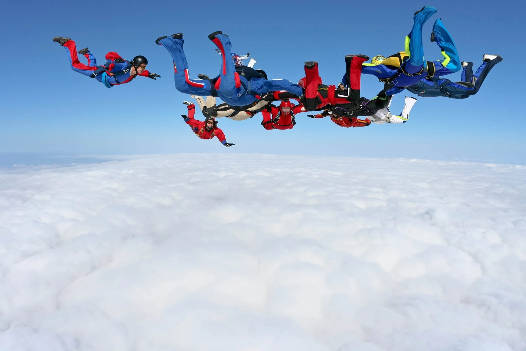 Group of skydivers in colorful jumpsuits performing aerial maneuvers above fluffy white clouds against a clear blue sky.