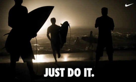Silhouettes of surfers on a beach at sunset, with the text "JUST DO IT" prominently displayed, illustrating Nike's inclusive and motivational branding message.