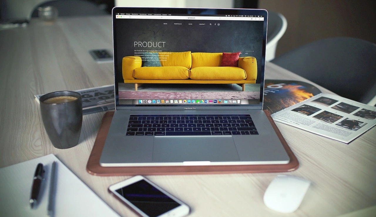 Laptop displaying a product page featuring a yellow sofa, with a coffee cup and smartphone on a desk, illustrating online consumer decision-making influenced by digital marketing.