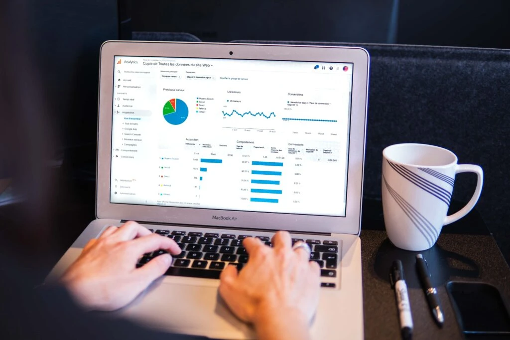 Person analyzing website analytics on a MacBook Air, displaying graphs and data metrics, with a coffee mug and pens nearby, emphasizing the importance of data in market research.