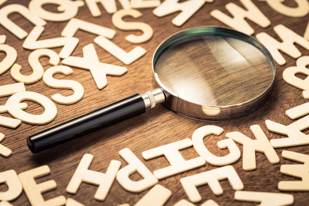 Magnifying glass on wooden surface surrounded by scattered alphabet letters, symbolizing exploration and understanding of marketing and market research terms.