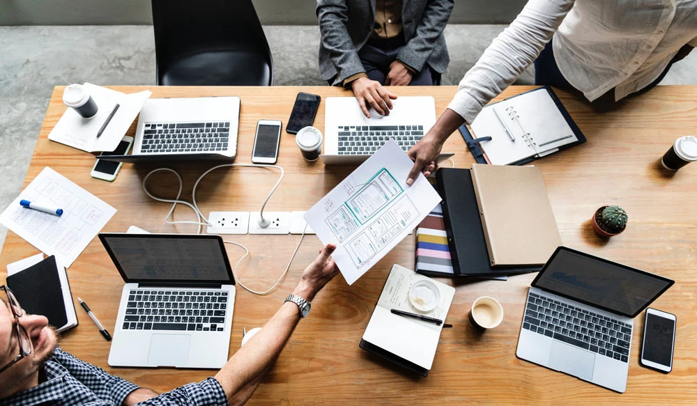 Group of professionals collaborating at a table with laptops, documents, and coffee, discussing packaging market research strategies and design optimization.