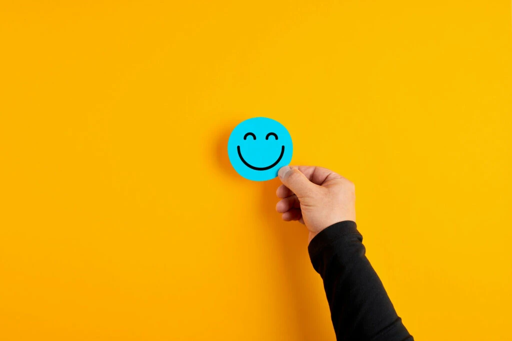 Hand holding a blue smiley face sticker against a bright yellow background, symbolizing customer satisfaction in the context of Net Promoter Score discussions.