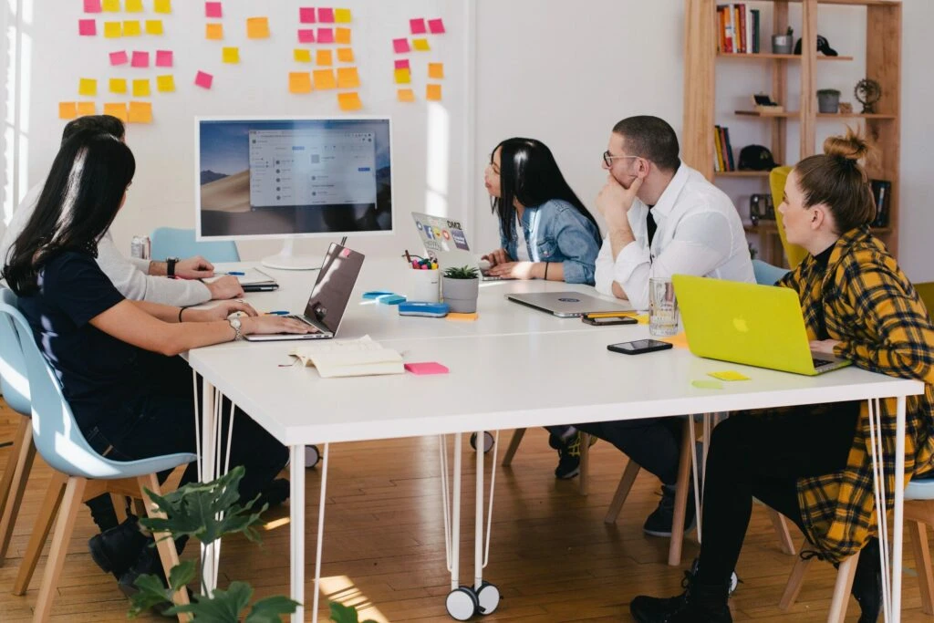 Group of diverse individuals engaged in a concept testing meeting, analyzing data on a computer screen, with laptops and notes on the table, surrounded by colorful sticky notes.