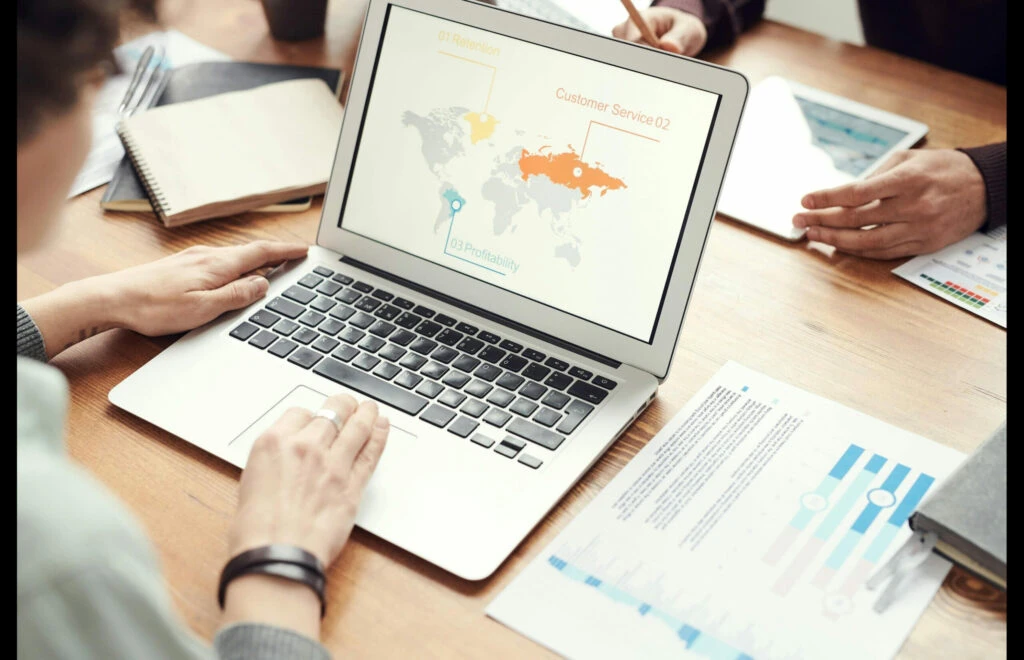 Person analyzing a laptop screen displaying a world map with customer service and profitability metrics, surrounded by documents and notes, illustrating brand tracking in crisis management.