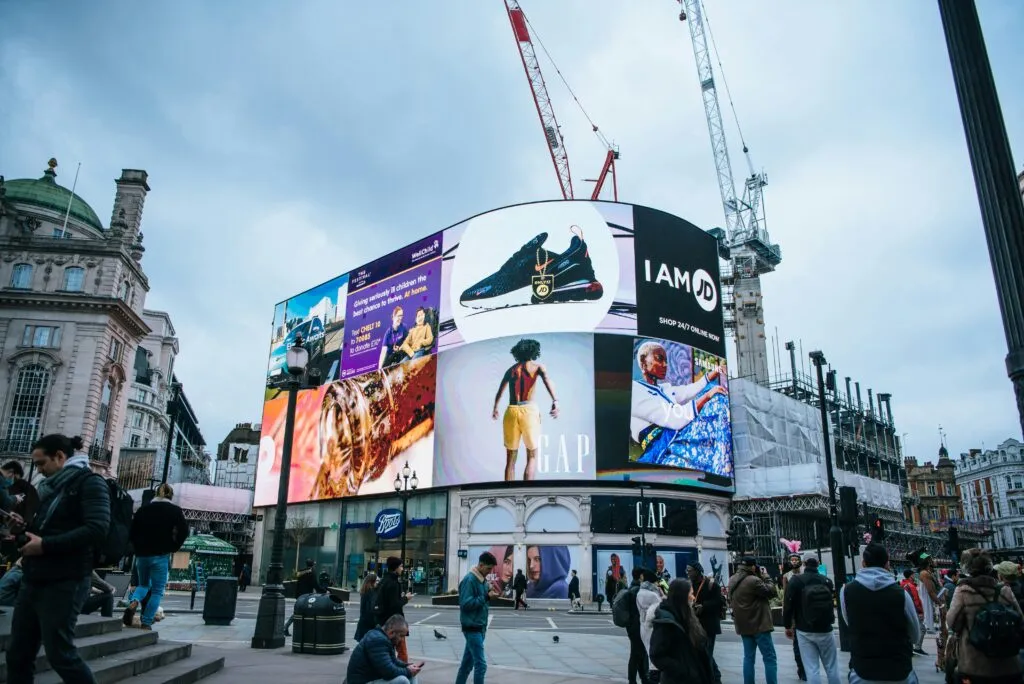 Outdoor advertising display in a busy urban setting featuring various brands and promotions, including sneakers, clothing, and lifestyle imagery, with pedestrians engaging nearby.